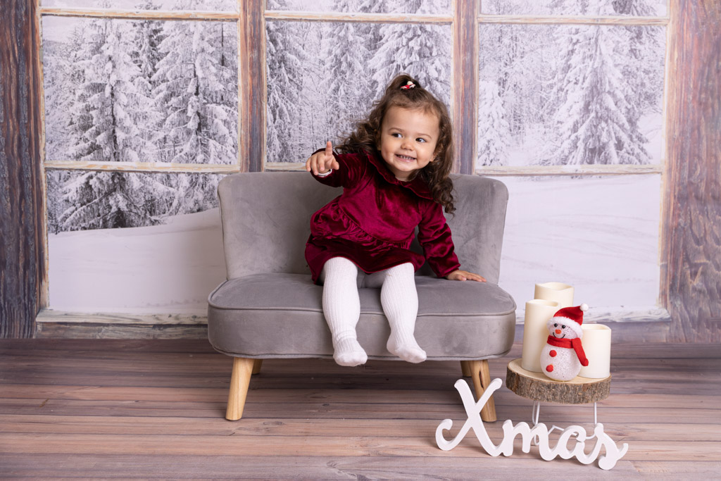 Kleine sMädchen sitzt auf einer Bank und lächelt, im Hintergrund ist eine Weihnachtslandschaft zu sehen