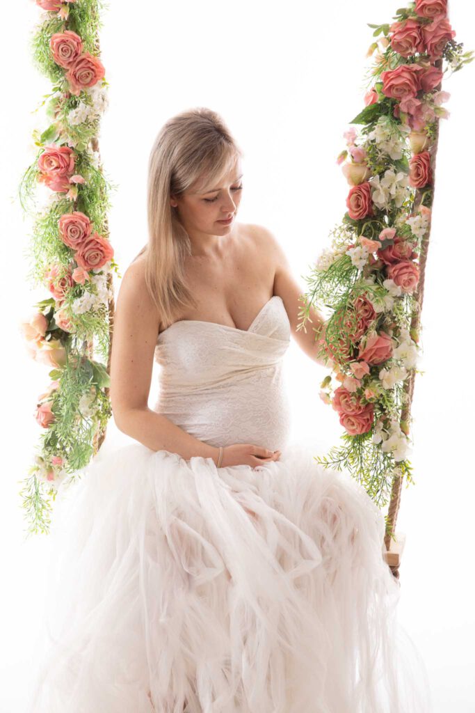 Schwangere in einem weißen Kleid sitzt auf einer Schaukel und schaut verträumt auf die Blumen