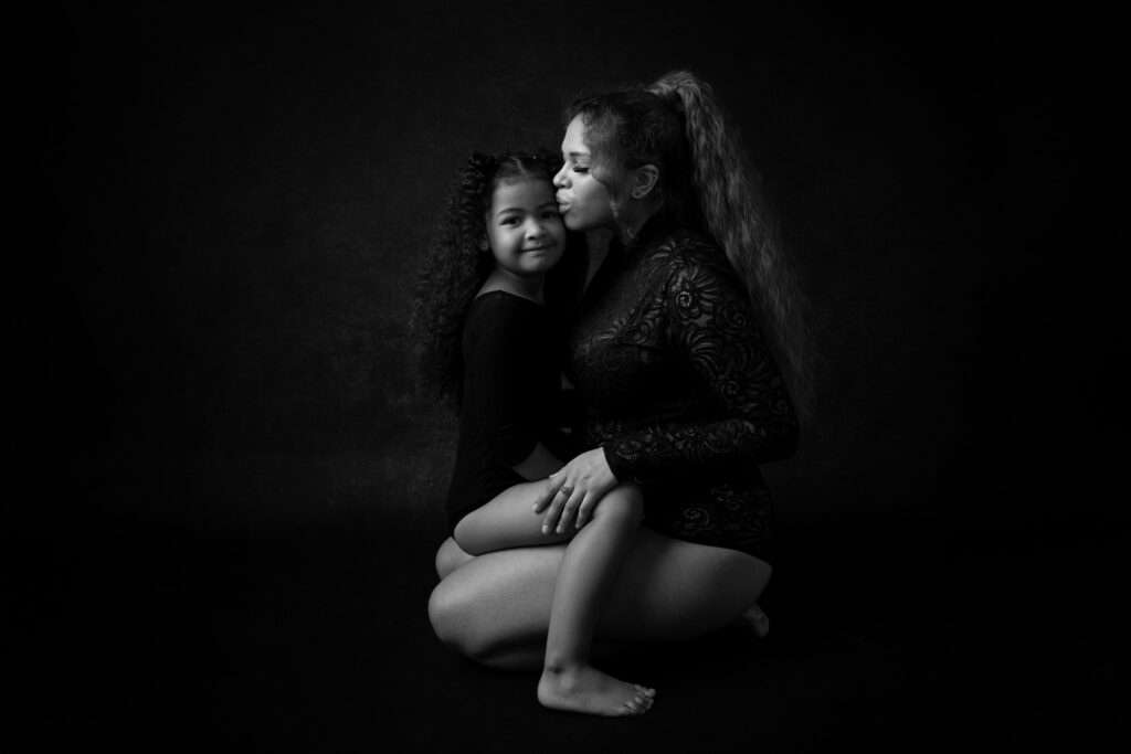 Baldmama sitzt mit ihrer Tochter auf dem Schoß auf dem Boden. Sie küsst liebevoll ihre Tochter