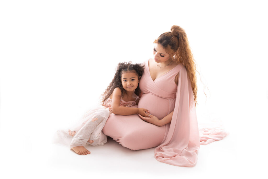 Schwangere sitzt neben ihrer Tochter auf dem Boden. beide tragen ein rosanes Kleid. fotostudio lohmar