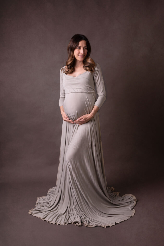 Schwangere vor einen dunklen Hintergrund in einem cremefarbenen Kleid, sie hat die hände liebevoll am Bauch