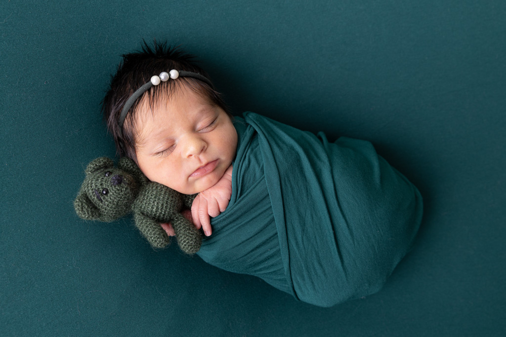Baby liegt eingewickelt in einer grünen Decke und hält einen Teddy in den Händen